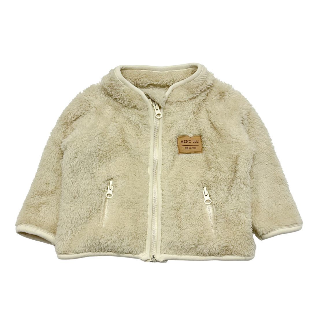 Chaqueta de pelo sintético color crema con cremallera, mangas cortas y un pequeño parche en el pecho.