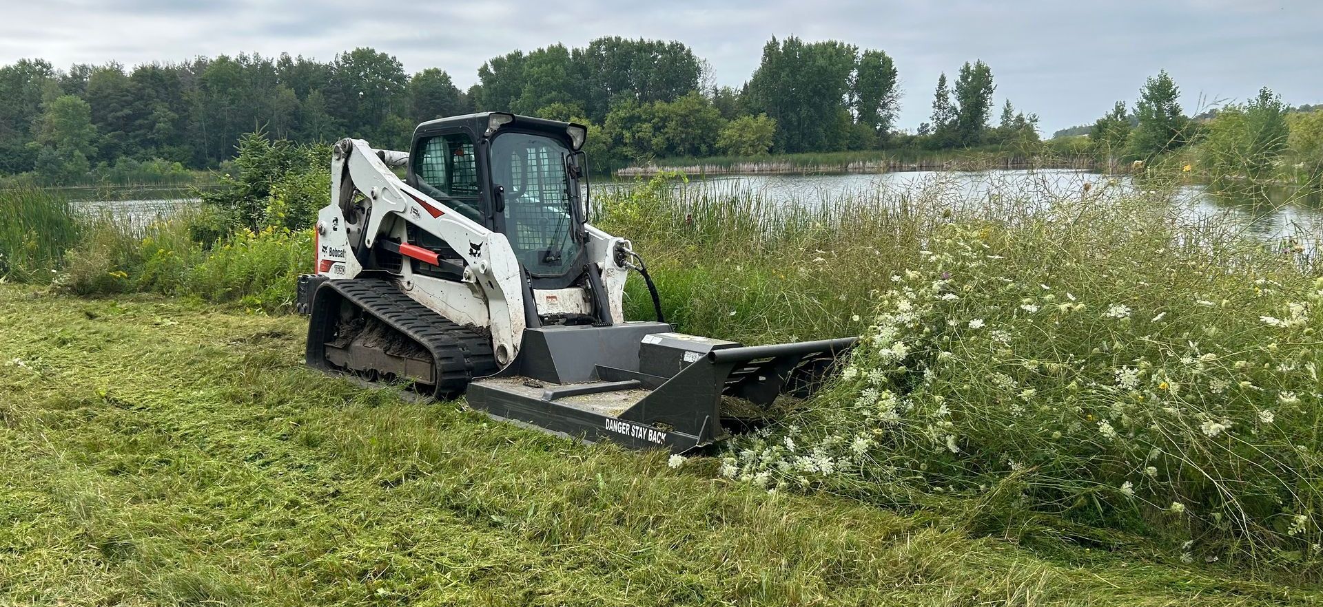 A bobcat is cutting grass in a field near a lake.