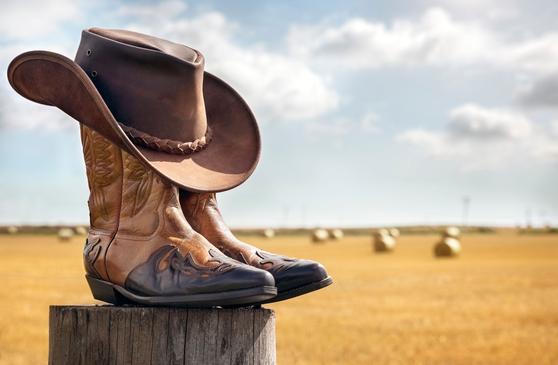 Cowboy boots and hat on wooden post in a field of hay bales under a cloudy sky.