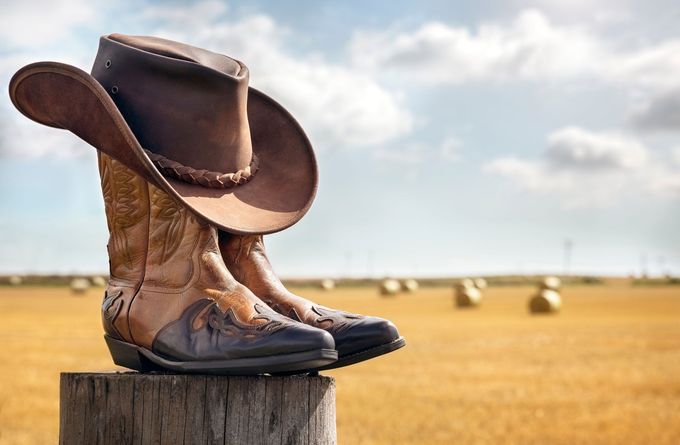 Cowboy boots and hat on wooden post in a field of hay bales under a cloudy sky.