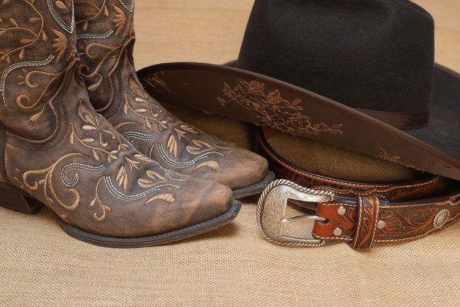 Cowboy boots, hat, and belt on a burlap surface. Brown, detailed leather with silver buckle.