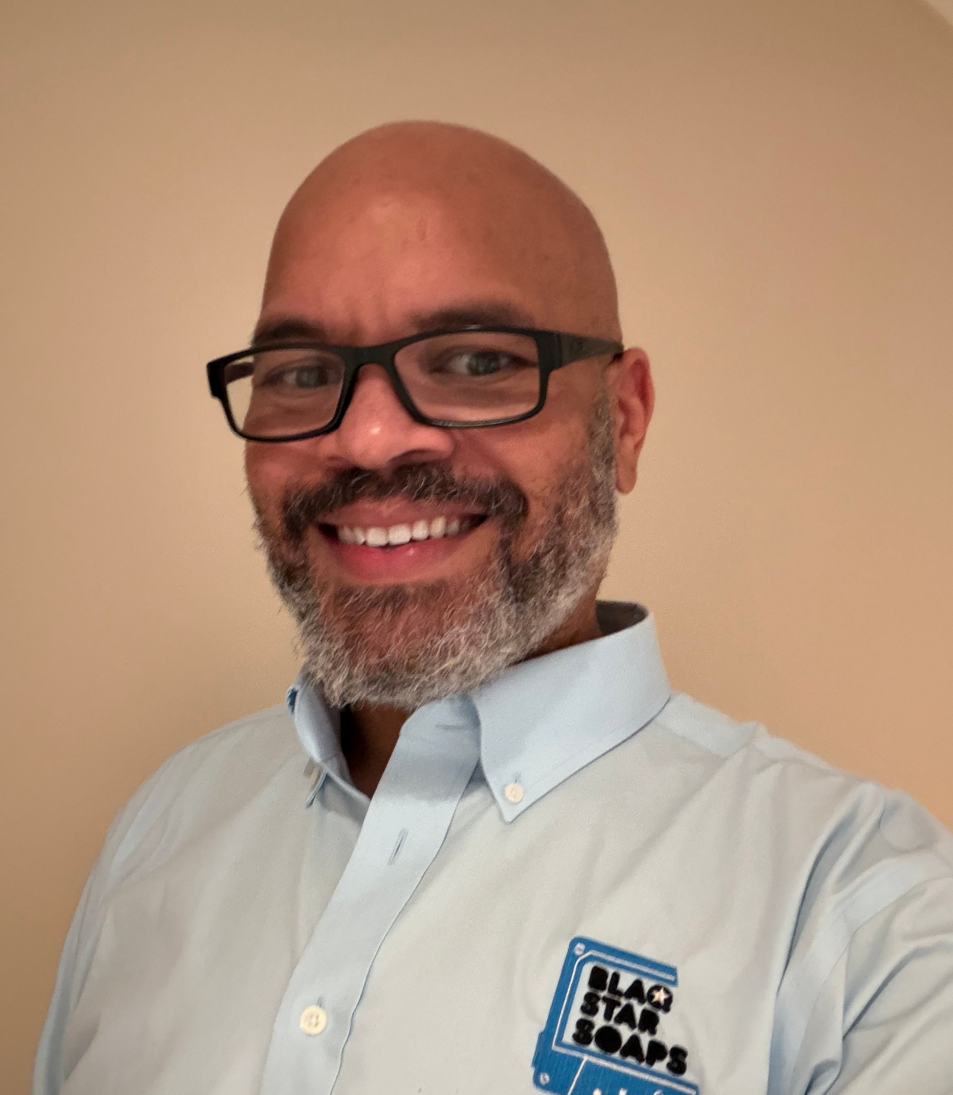 Person smiling in front of a transgender flag. They wear glasses and a striped shirt.