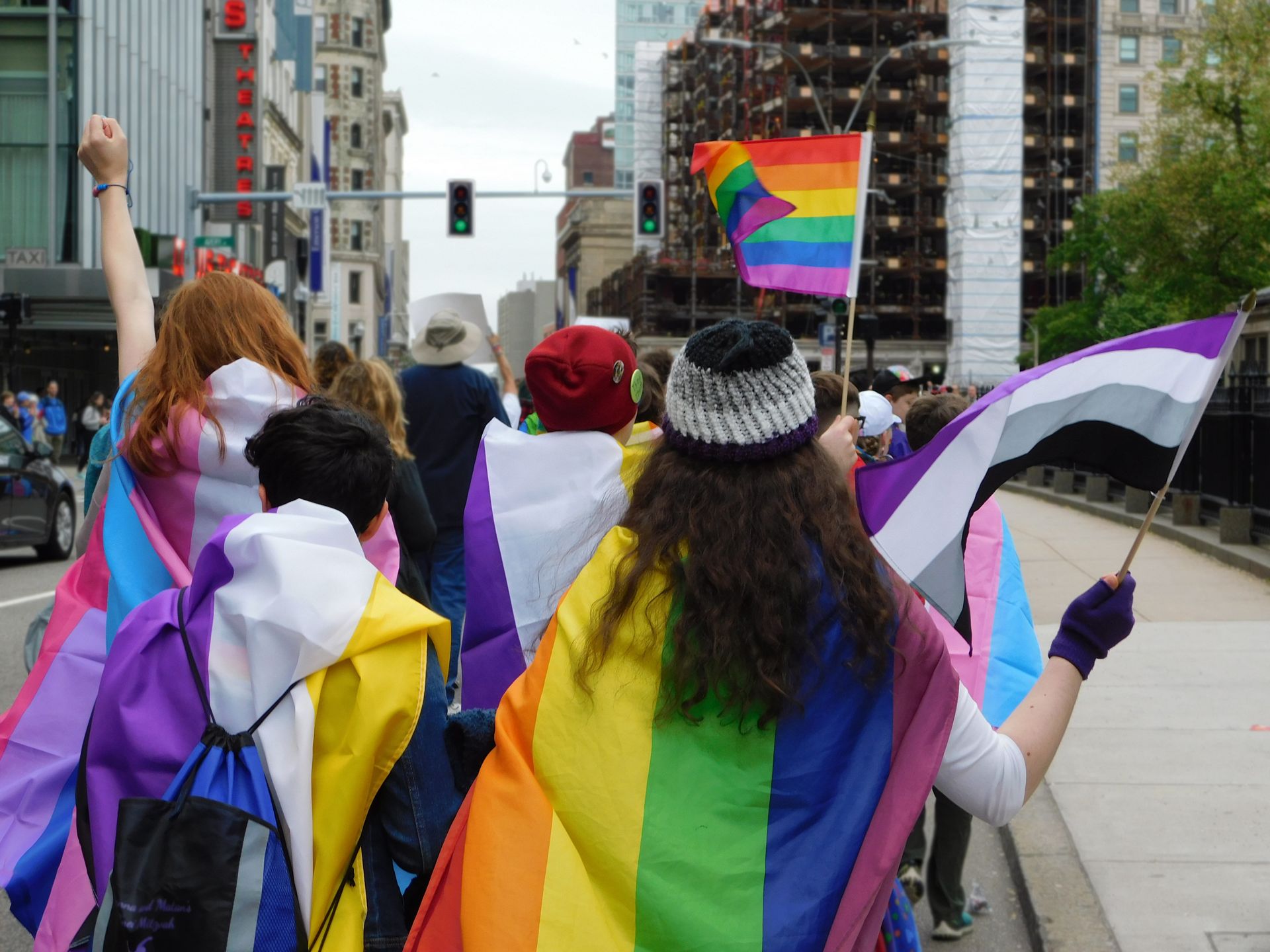 People in a pride parade, holding flags and wearing rainbow capes. Urban street setting.