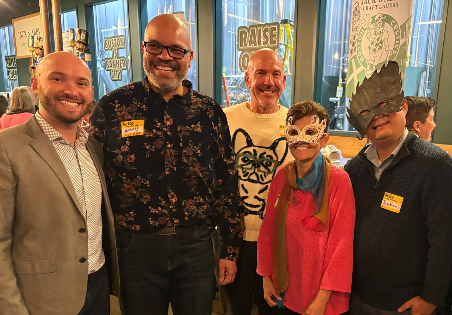 Group of six people smiling at a gathering, one wearing a mask, inside a building with a brewery backdrop.