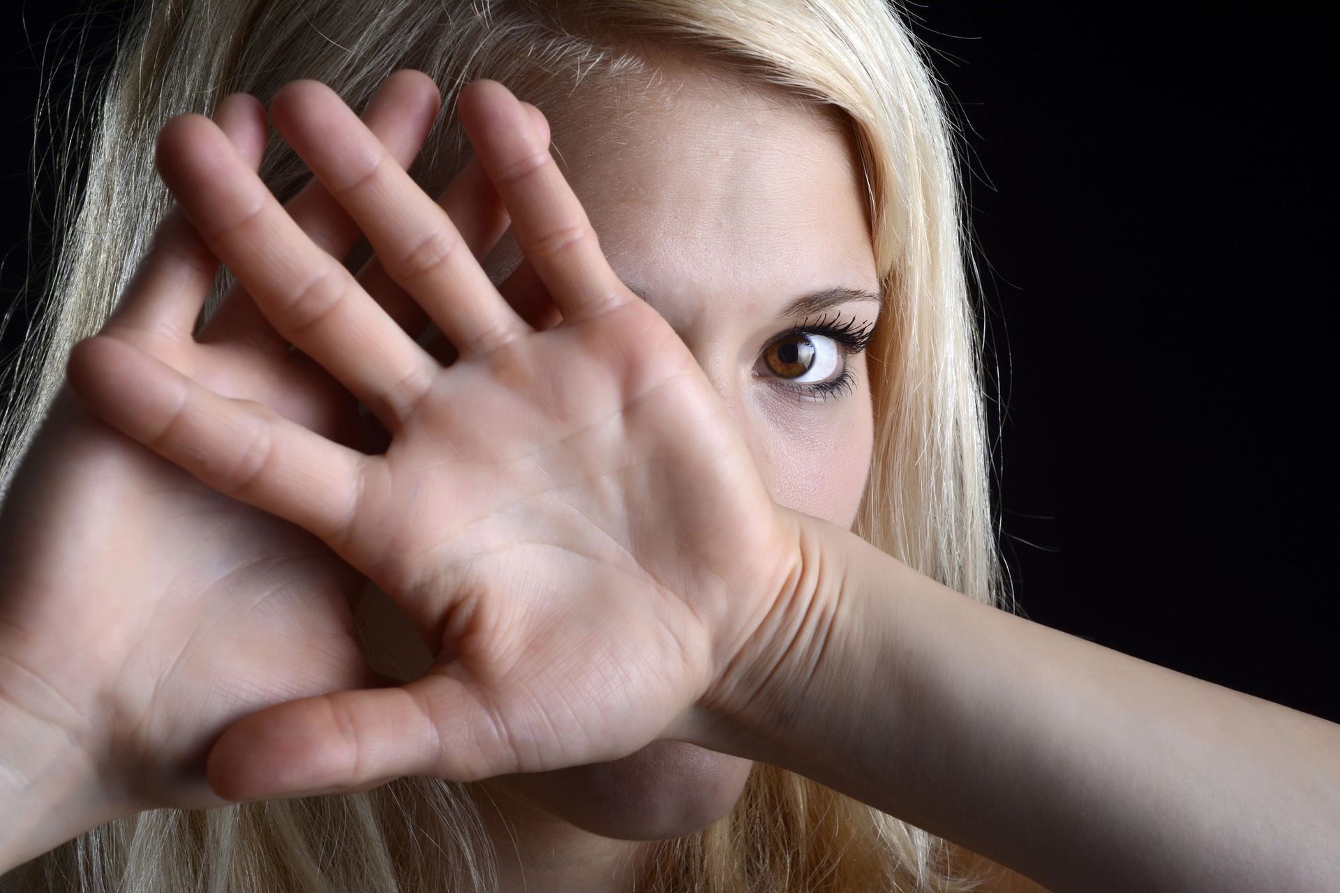 Blond woman holding hands up, partially covering her face, looking at viewer.