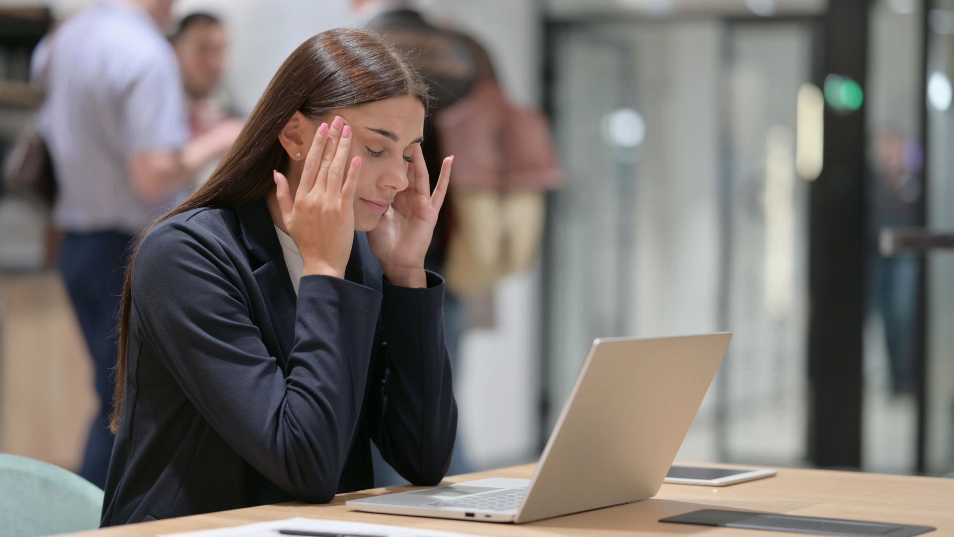 Een vrouw met gesloten ogen en haar handen voor haar slapen, werkt op een laptop aan een tafel op kantoor.