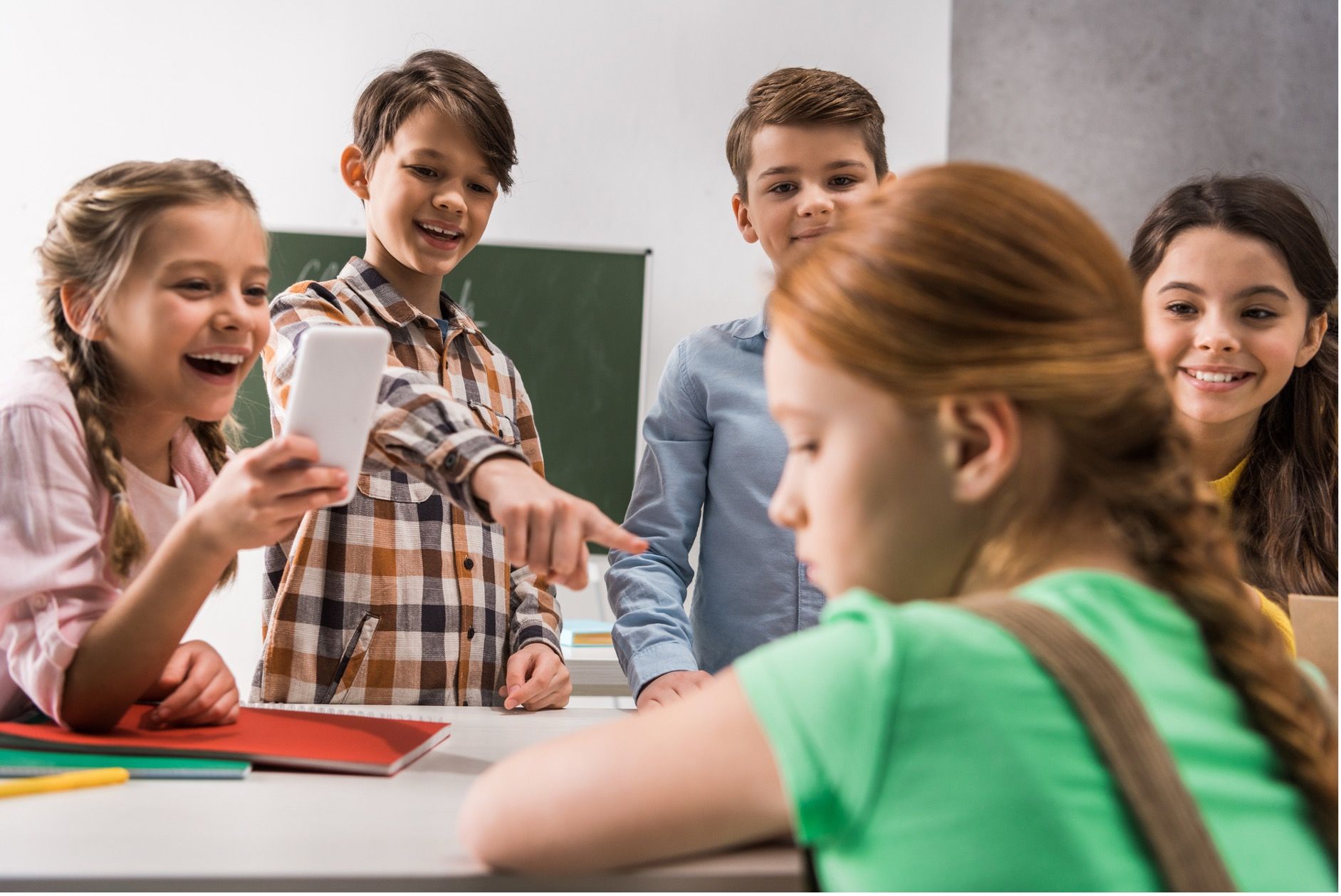 Group of children laughing, pointing at a student. One holds a phone. Classroom setting.