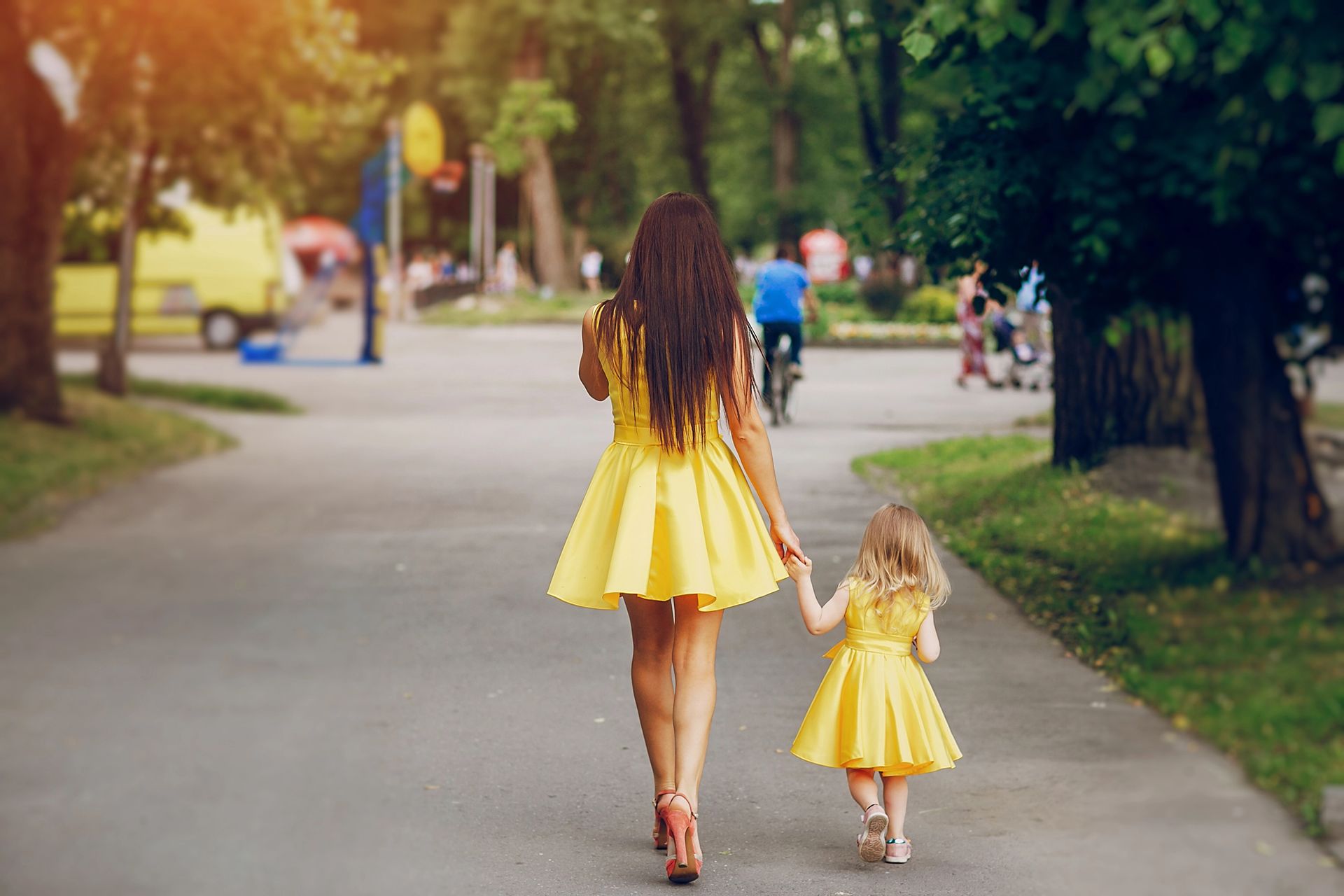 Vrouw en kind in bijpassende gele jurken houden elkaars hand vast terwijl ze over een pad in een park lopen.