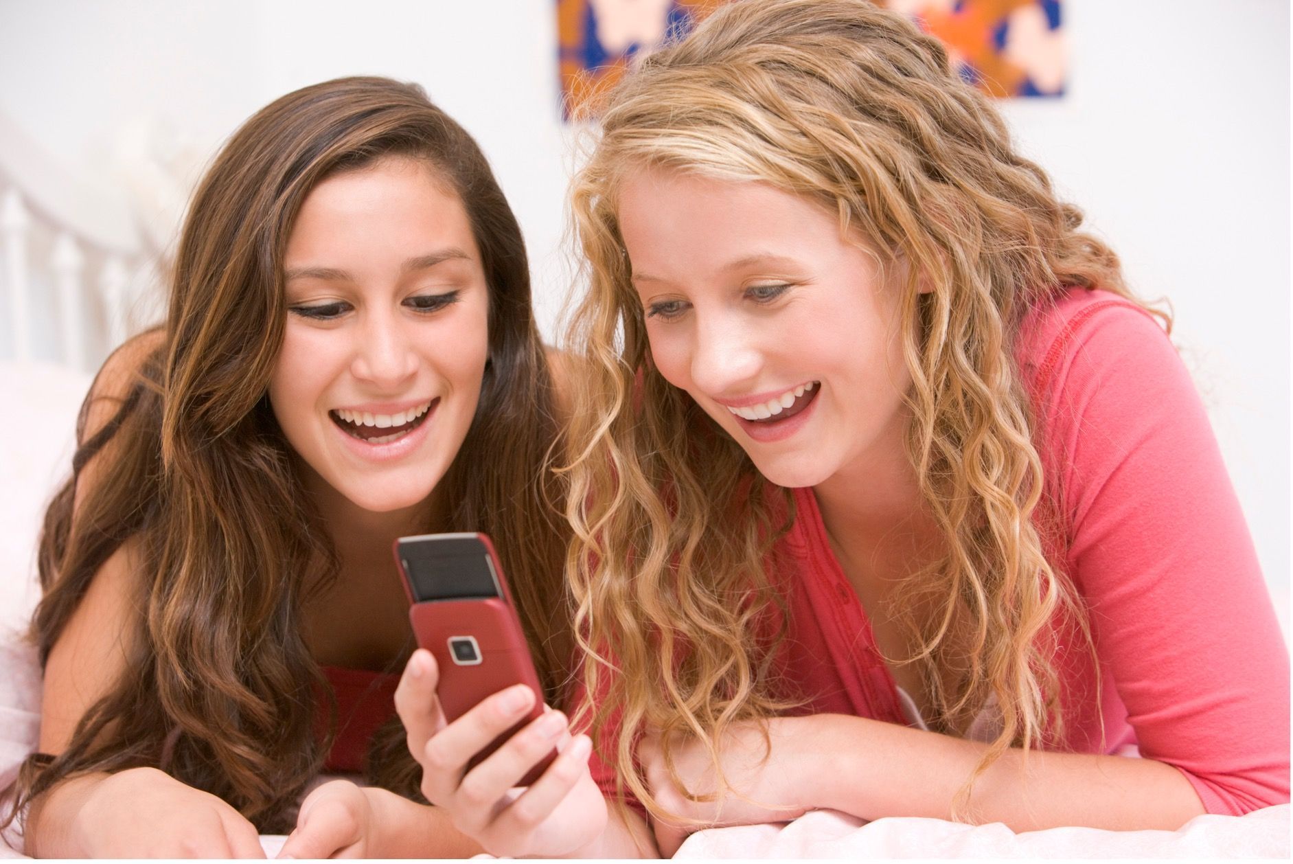Two young women, smiling, looking at a cell phone, lying on a bed.