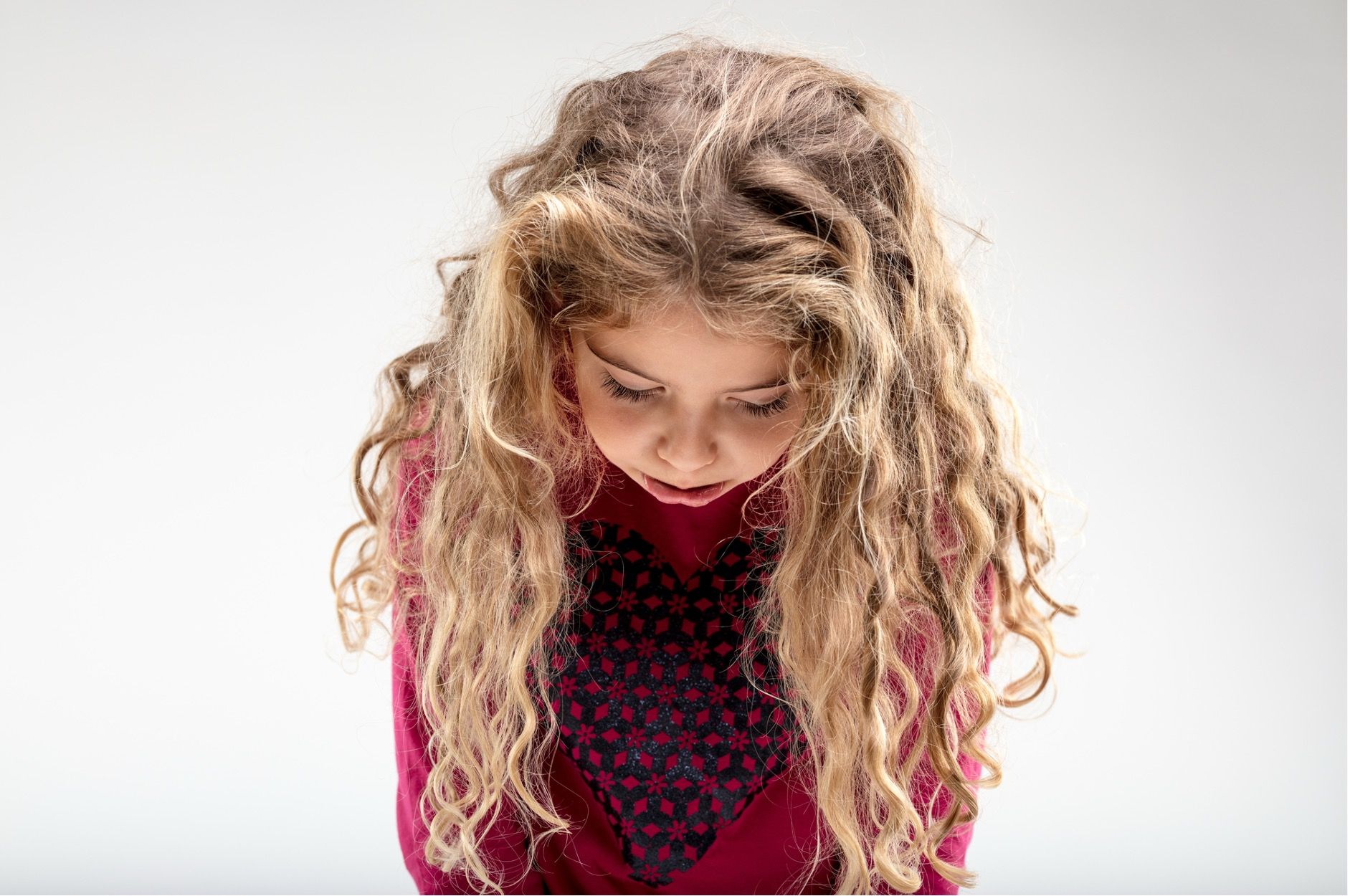 Meisje met lang, golvend bruin haar kijkt naar beneden. Ze draagt ​​een rode trui tegen een witte achtergrond.
