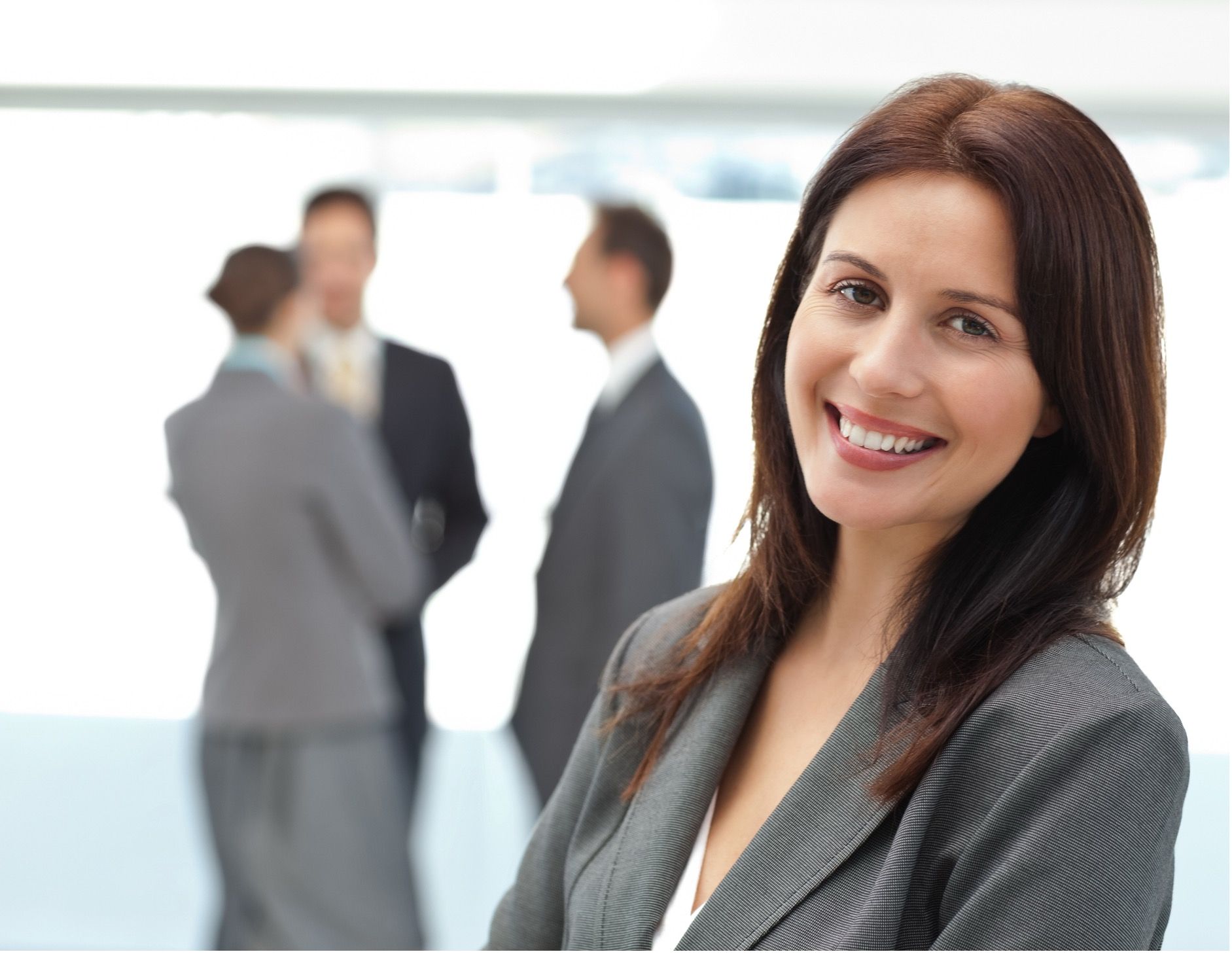 Woman in business attire smiles, arms crossed, with colleagues blurred in the background.