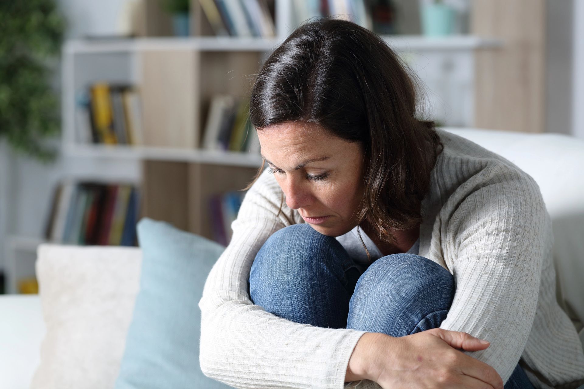 Vrouw zit op de bank, hoofd gebogen, knieën op haar borst, en ziet er verdrietig uit.
