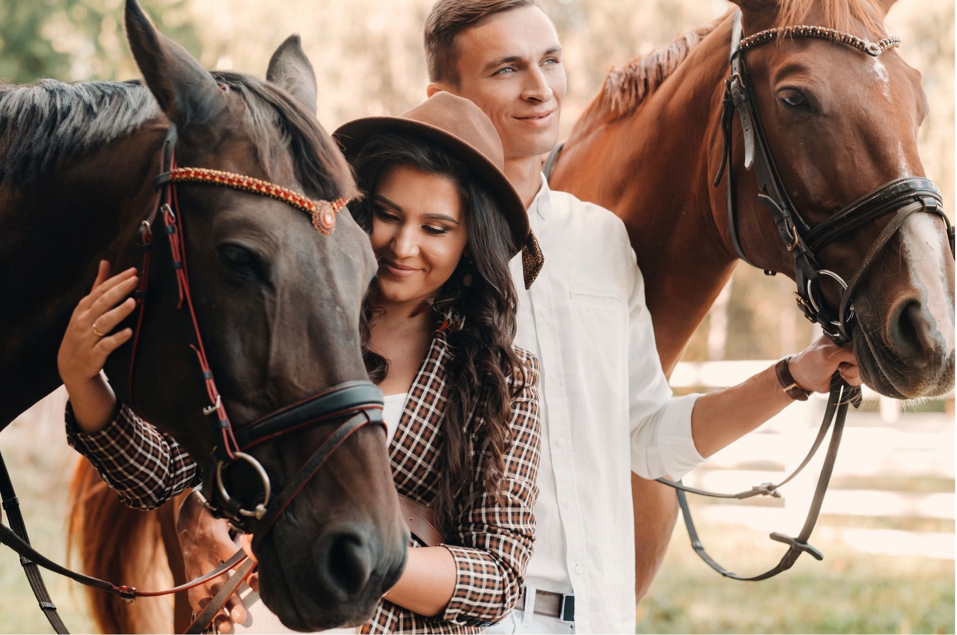 Couple petting horses. Woman in hat, man smiles. Outdoors, sunny day.