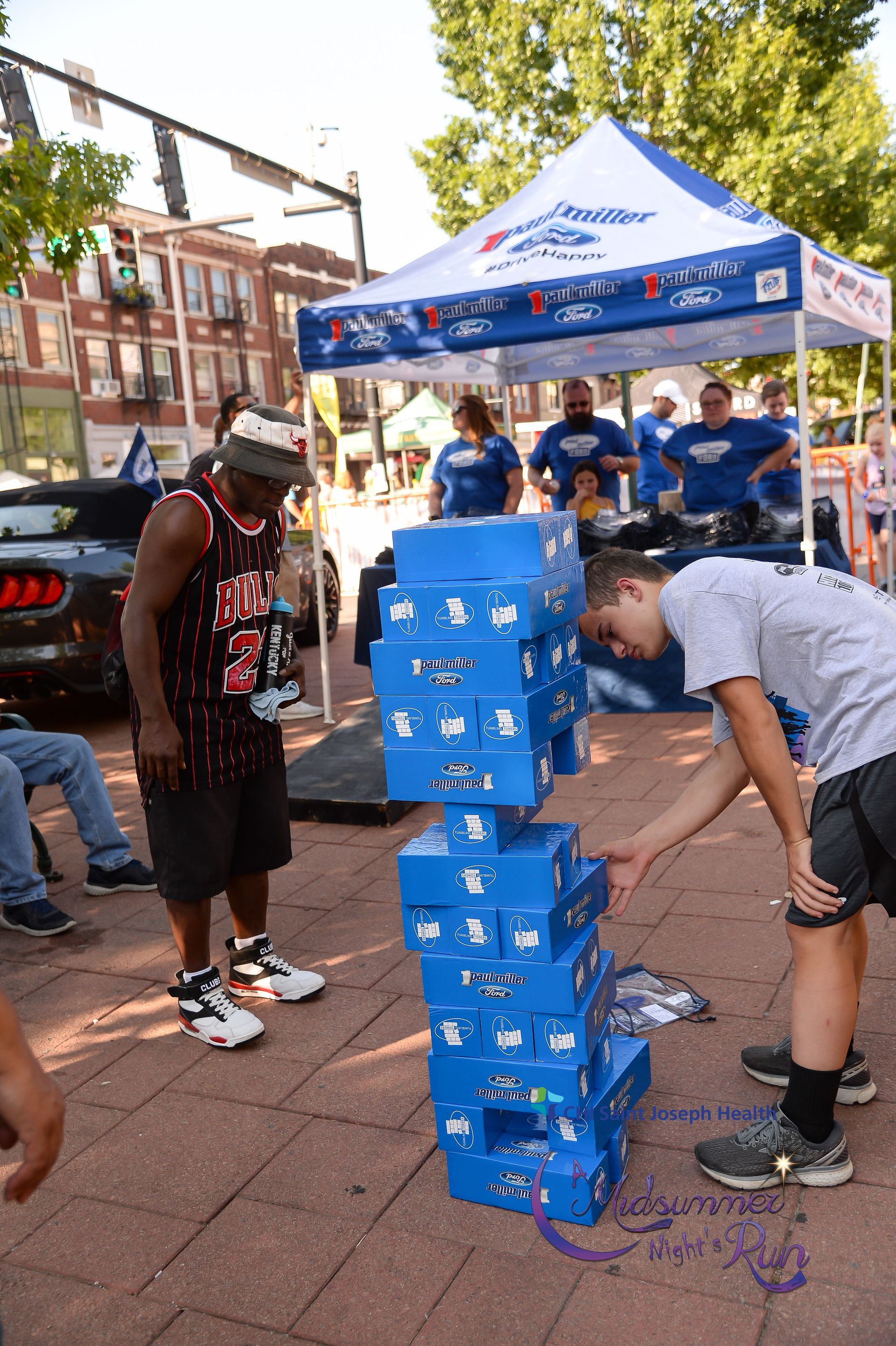 Man and boy stacking blue boxes outside under a tent. Others watch in the background.