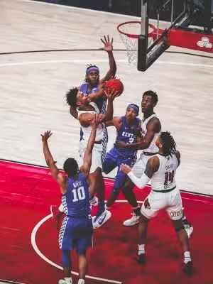 Basketball players in action, one shooting with several others blocking, on a red court, under the hoop.
