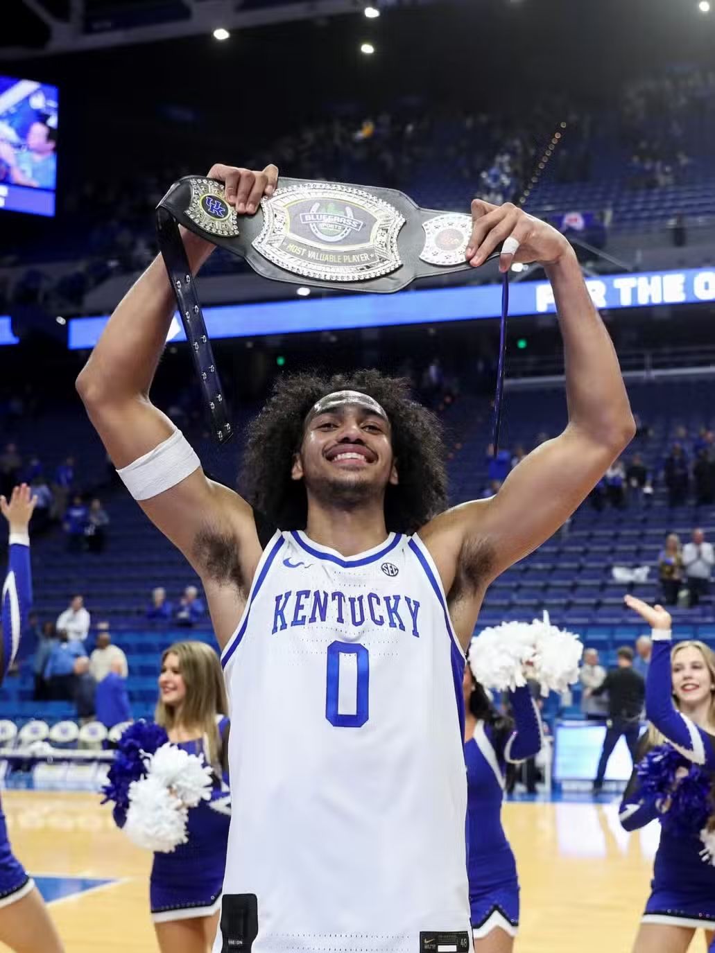Basketball player in a Kentucky jersey holds a championship belt over his head, celebrating in a crowded arena.