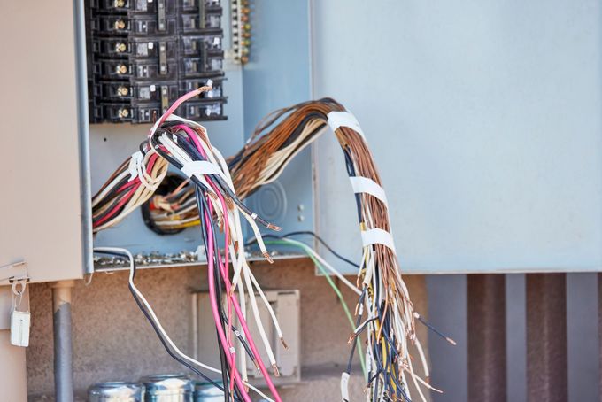 A wall-mounted electrical panel with many exposed, multicolored wires hanging loose and disconnected.