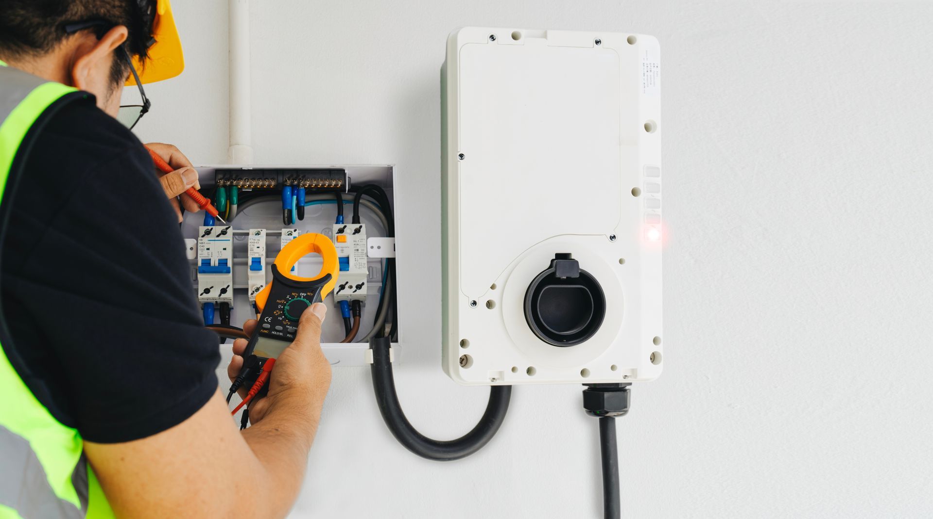 An electrician in a high-visibility vest uses a clamp meter to test electrical components inside a wall-mounted EV charger.