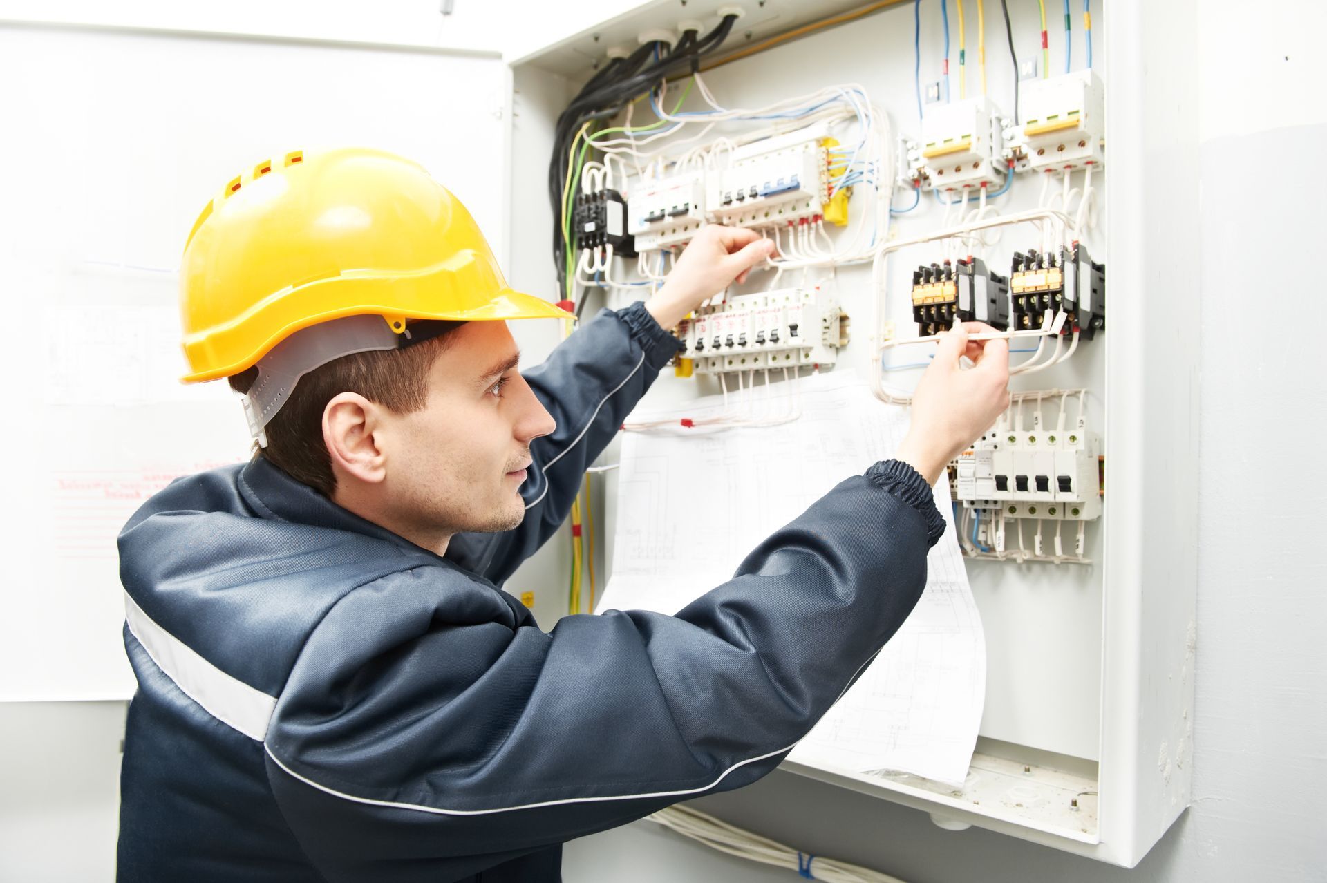 Electrician in a yellow hard hat and uniform working on wires inside an open electrical distribution panel.