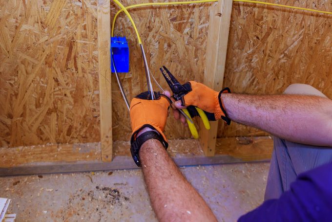 A person wearing orange work gloves uses pliers to strip insulation from yellow electrical wire at a wall-mounted blue box.
