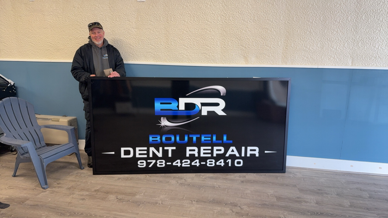 Man stands next to a BDR Boutell Dent Repair sign. Black and blue sign, outside.