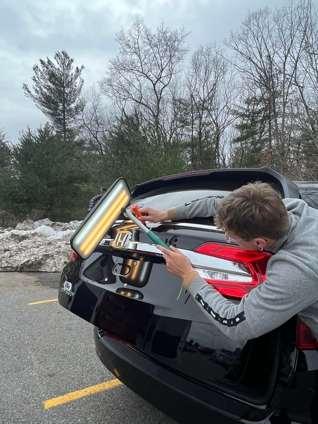 Person using dent removal tools on a black car's trunk, outdoors near snow and trees.