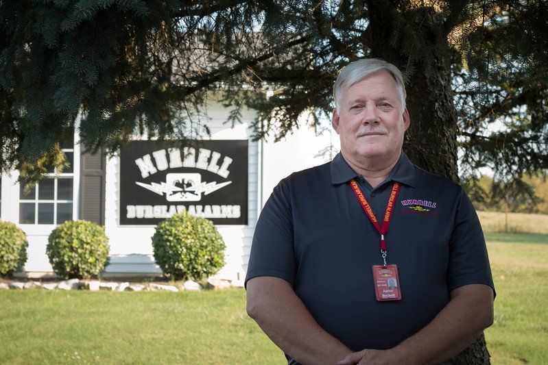 Man standing in front of a school building sign, wearing a dark polo and red lanyard, outdoors on a sunny day