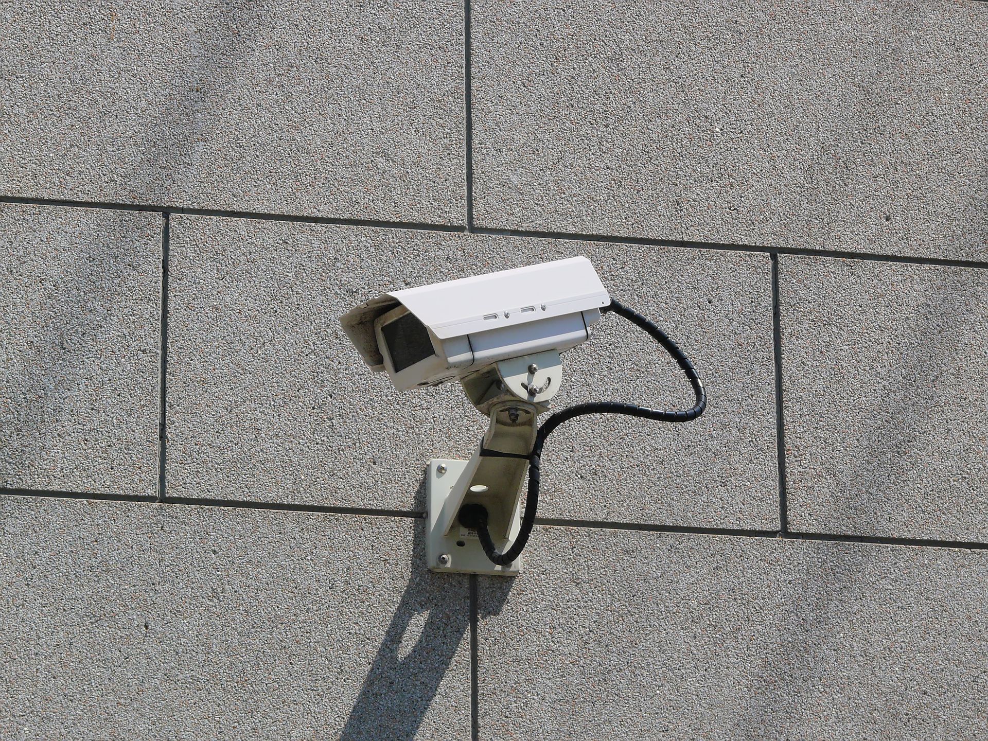 Security camera mounted on a gray, stone-tiled building, aimed downwards. Security camera mounted on a gray, stone-tiled building, aimed downwards.