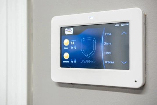 Wall-mounted touchscreen security control panel with blue interface on a white wall