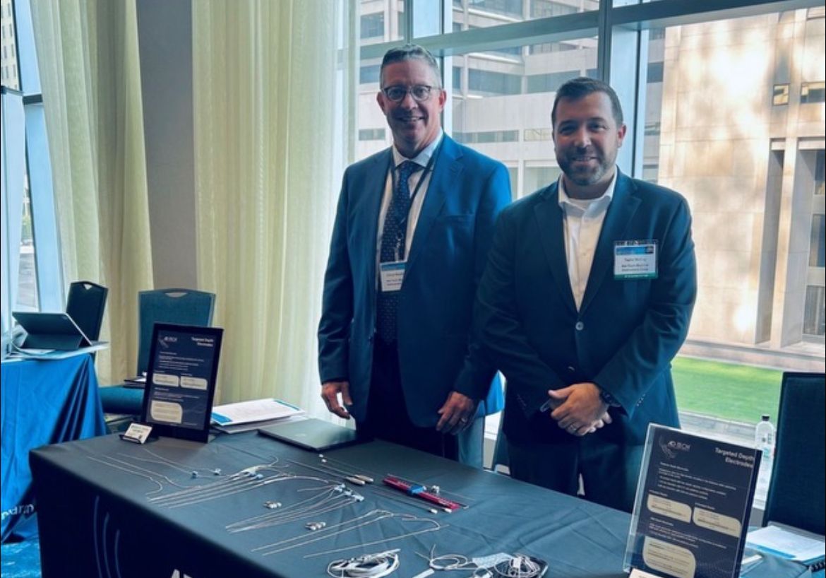 Two men at a booth, likely a conference. They are smiling, standing by a table with brochures, in a well-lit room.