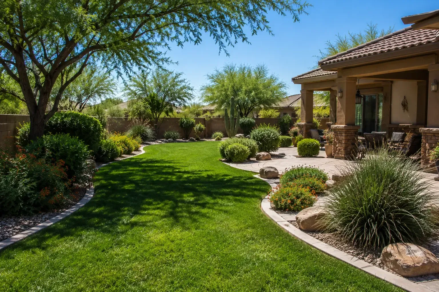 Sunlit landscaped backyard with green lawn, shrubs, trees, and a stucco patio house