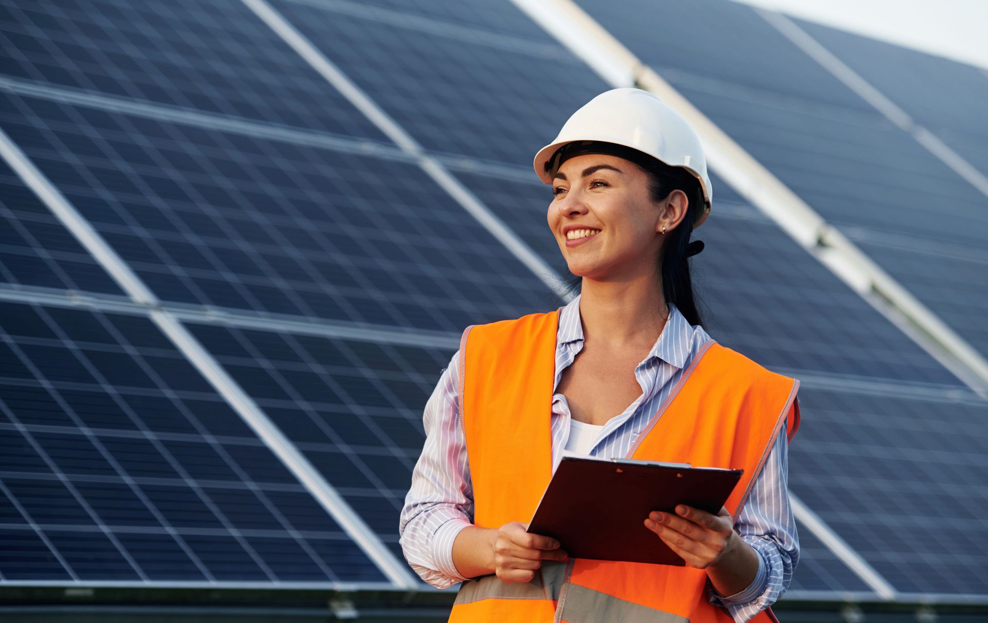A woman is standing in front of a solar panel holding a clipboard.