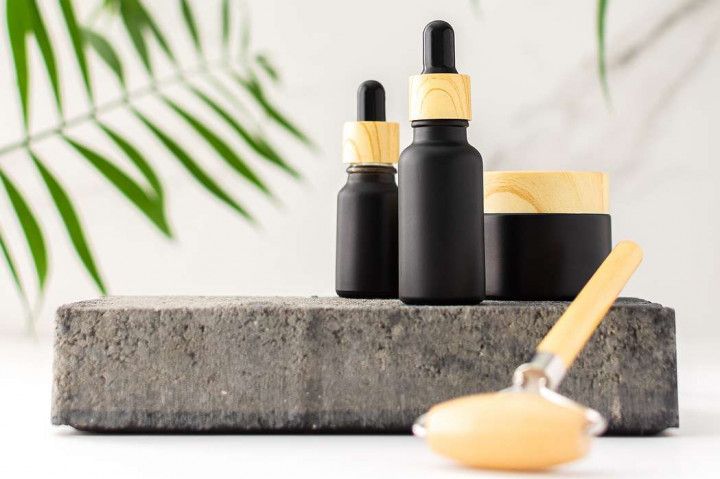 Black skincare bottles with gold caps on a concrete block, with a cream jar and dropper in front.