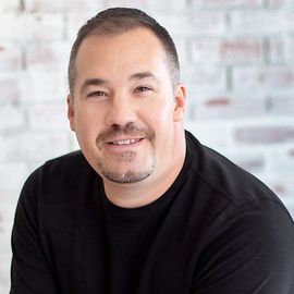 Smiling man in a black shirt posing against a light brick wall background