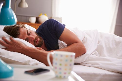 Person sleeping in bed with white blanket, next to a colorful mug and smartphone on the nightstand.