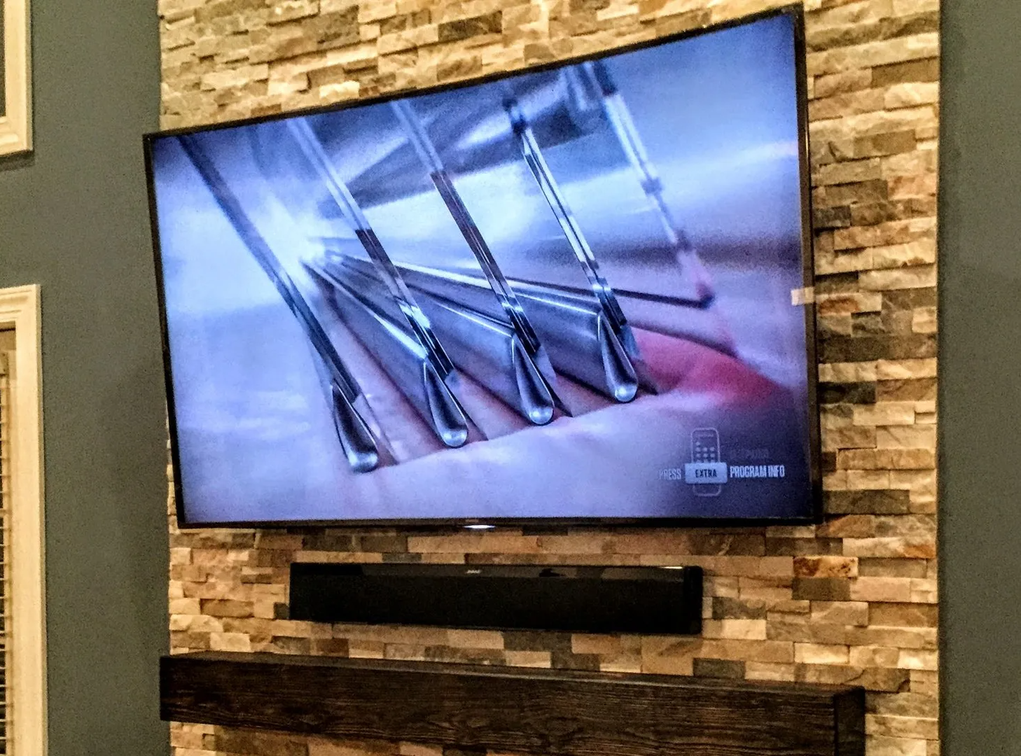 Large curved TV on a stone wall, with a sound bar and dark wooden mantle below. TV is on.