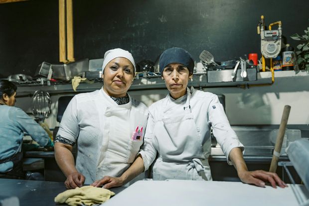 Dos chefs con uniformes blancos están de pie en una cocina industrial, uno de ellos con una mano apoyada en la encimera.