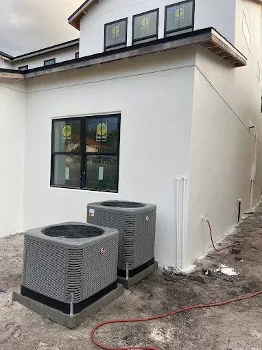 Two air conditioning units near a white building with black framed windows. Construction site.