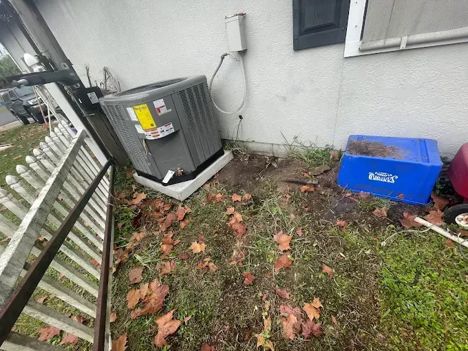 An air conditioning unit next to a house wall. Ground covered in leaves, a blue storage box sits nearby.