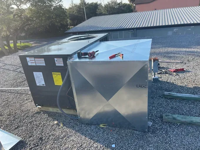 HVAC units on a rooftop. One is black, the other is silver. Tools are on top of the silver unit. Gravel ground.