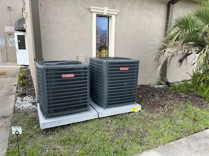 Two dark-colored Goodman AC units sit on concrete pads outside a building with a window and door.