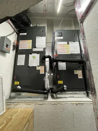 Two dark HVAC units in a white walled utility room. Black piping connects them. Light strip above.