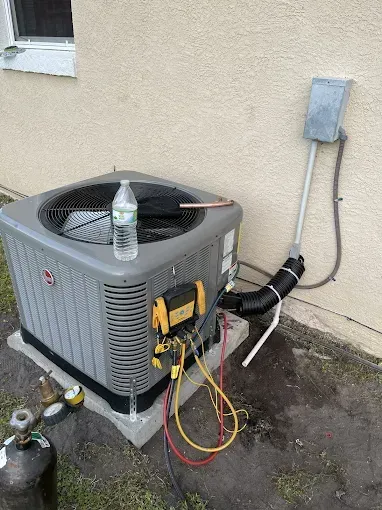 Air conditioner unit being serviced outdoors; attached hoses, water bottle on top.