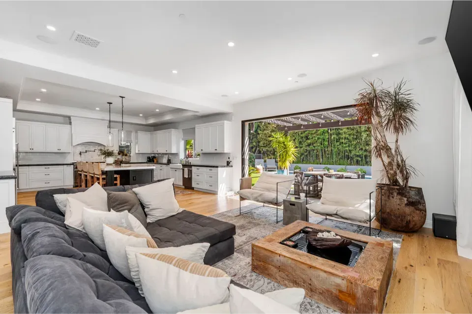 An open-concept home living space with a large grey sectional sofa and a view into a white kitchen and outdoor patio.