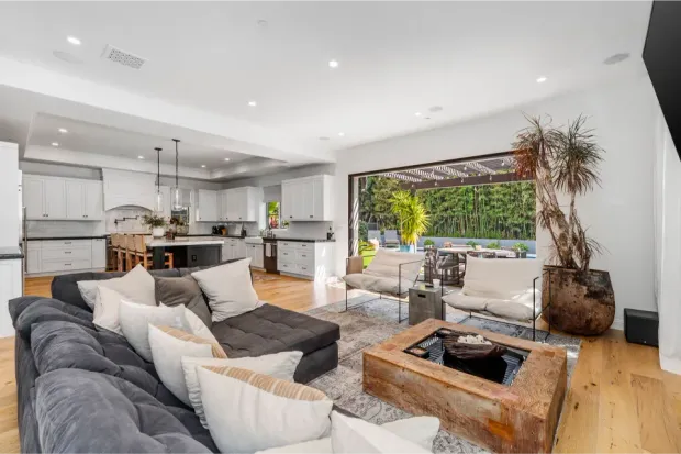 An open-concept home living space with a large grey sectional sofa and a view into a white kitchen and outdoor patio.