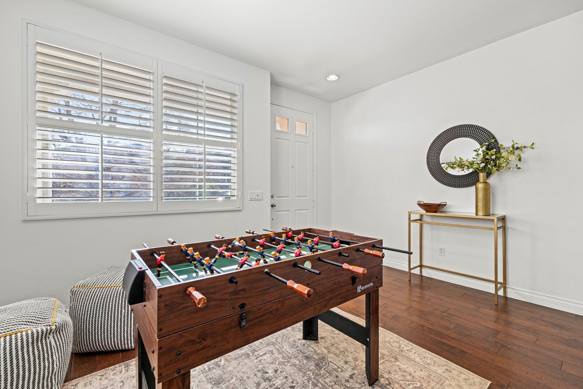 Sunlit interior flex space featuring a dark wood foosball table, patterned stools, and a gold console table.