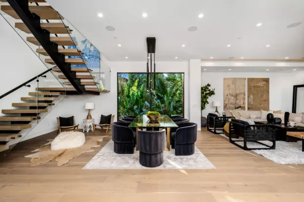 A spacious home dining area featuring a floating wooden staircase, a long gold table, and a view of tropical greenery.