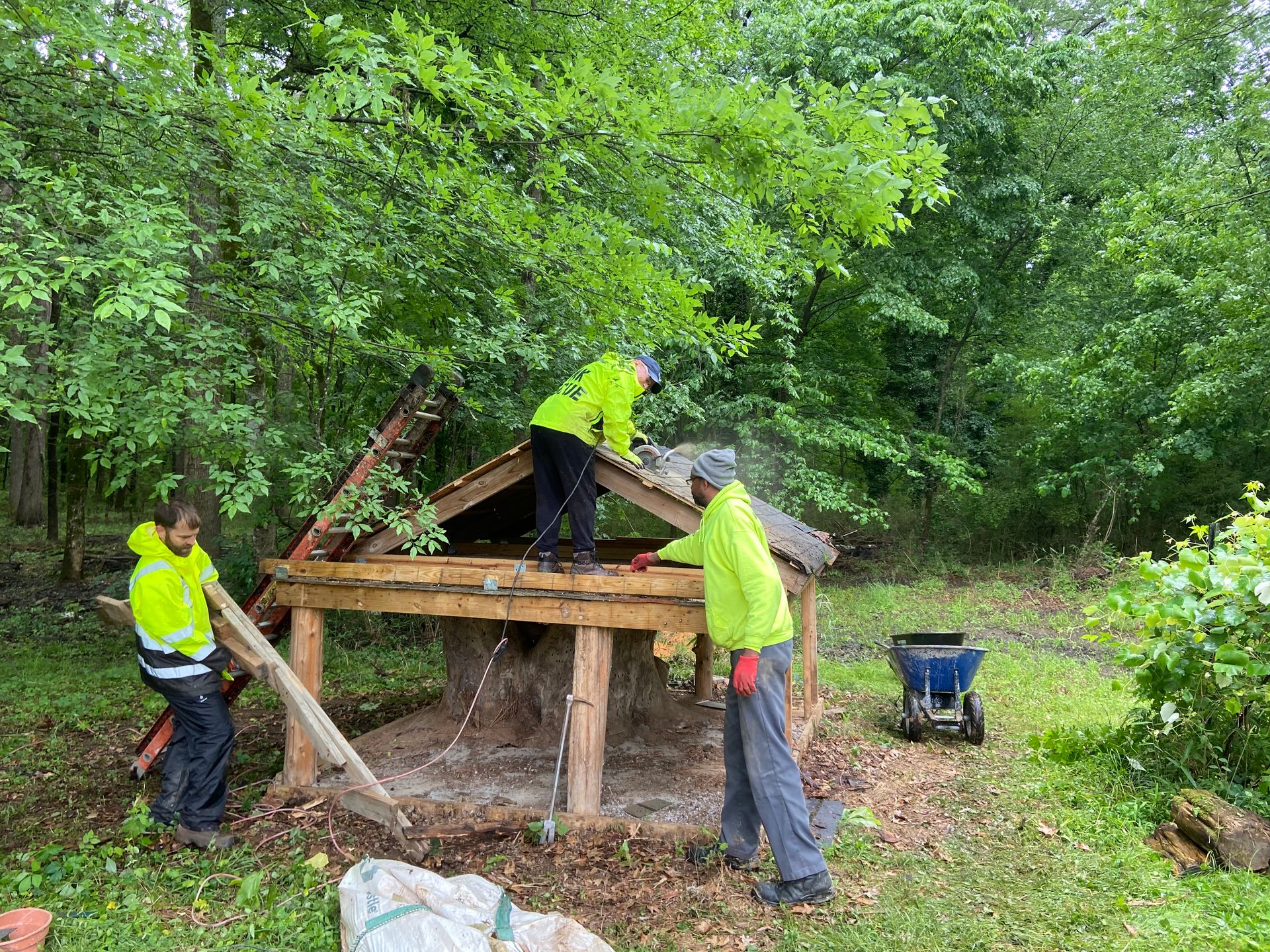 People in reflective vests repairing a wooden structure in a wooded area.