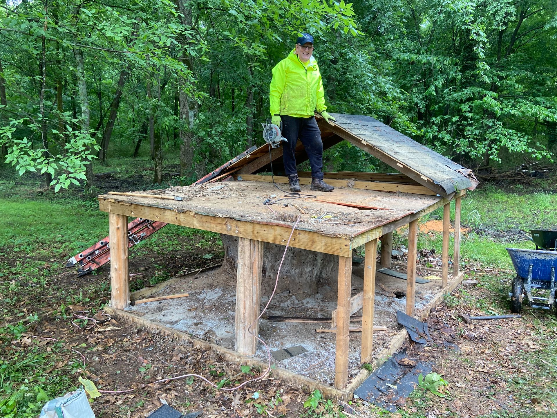 Person standing on a partially demolished wooden structure in a wooded area; wearing a yellow jacket.