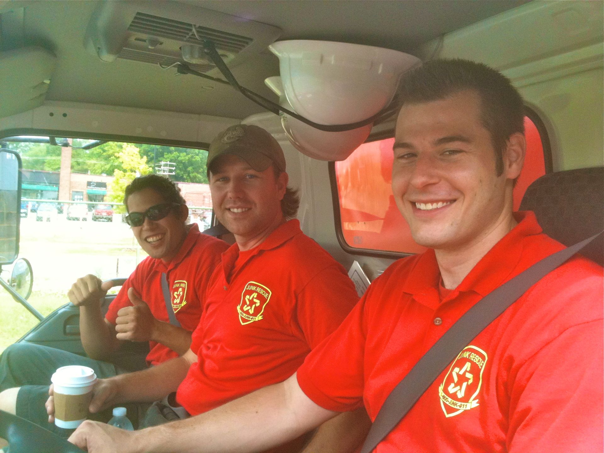 Three smiling people in red shirts inside a vehicle, possibly an ambulance. One gives a thumbs up.